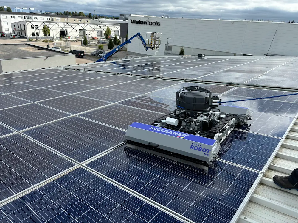 Robosun_Gallery_10 - Robosun Robotic solar panel cleaner on a roof sweeping across photovoltaic modules with a lift and industrial complex in the background.