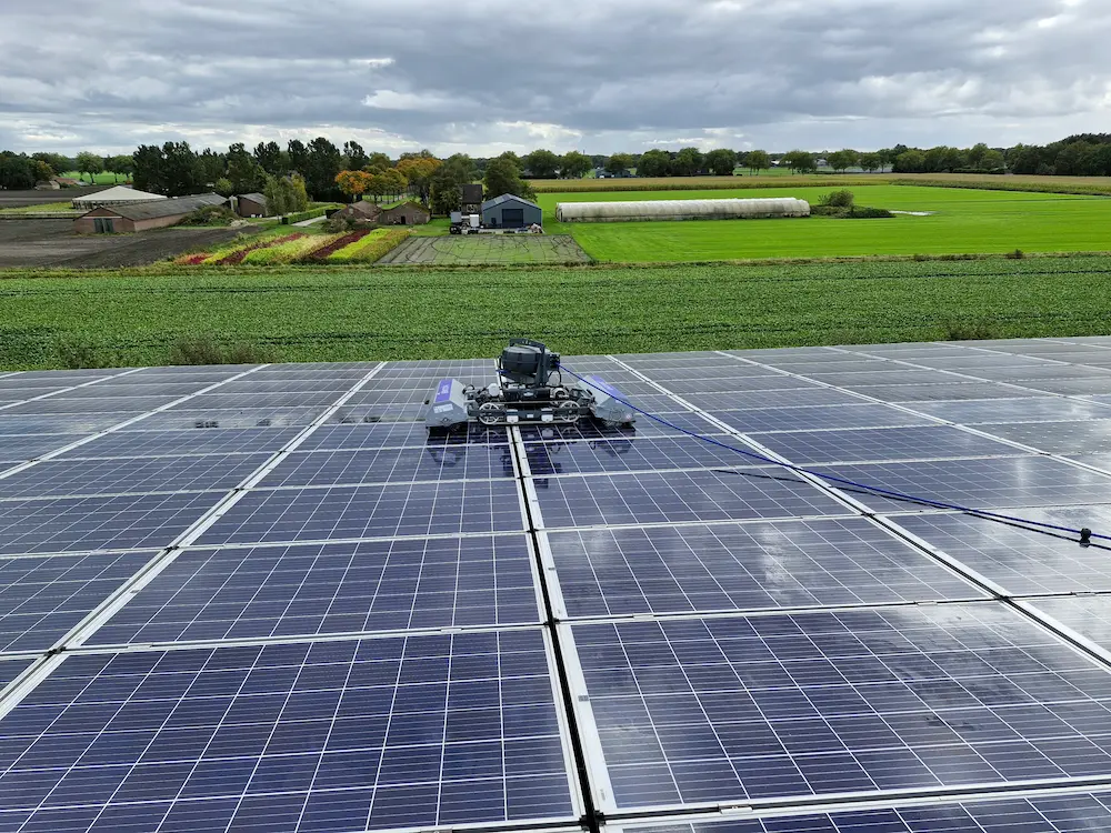Robosun_Gallery_13 - Robosun Robotic maintenance rover on a large solar panel field with farm fields and buildings in the distance under a cloudy sky, showing a solar farm setup.