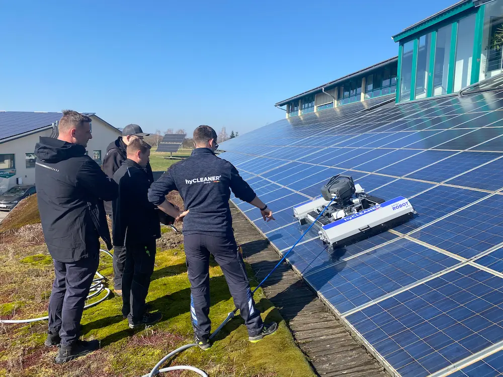 Robosun_Gallery_3 - Robosun Four workers stand on a sloped roof inspecting a robotic solar panel cleaner on the panels outside a building site.