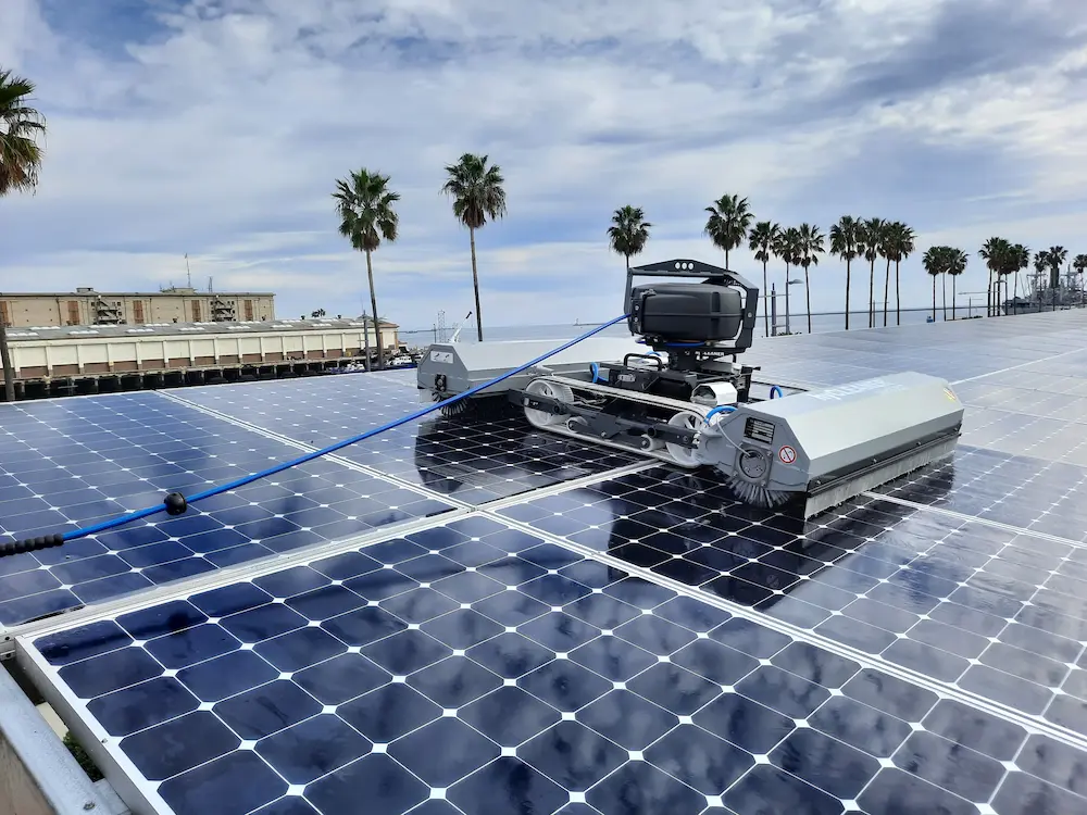 Robosun_Gallery_5 - Robosun Robotic solar panel cleaner moving over a flat roof covered in blue solar panels near a coastal area with palm trees in the distance.