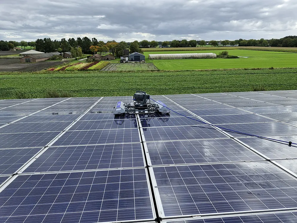 Robot-equipped maintenance vehicle on a large solar panel farm with green fields and farm buildings in the distance.